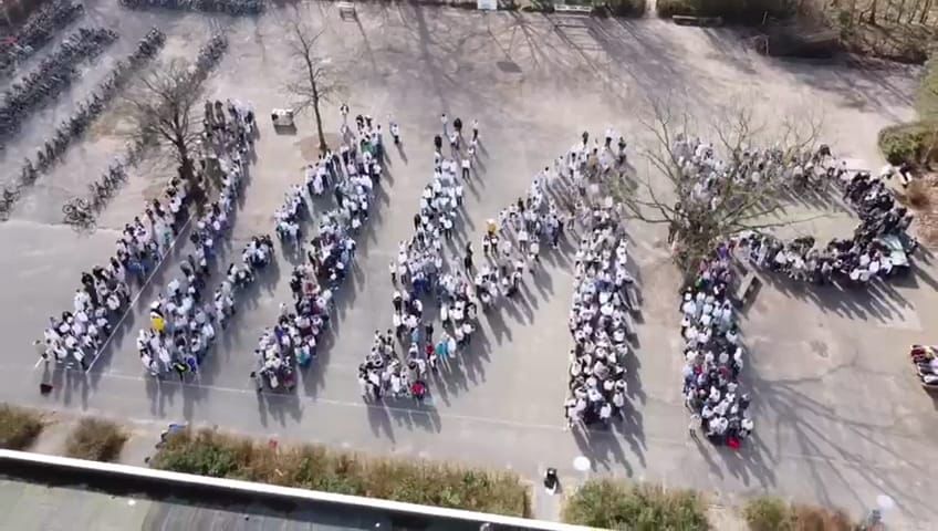 Menschen stehen auf einer Fläche und stellen die Buchstaben M N und P dar, viele tragen helle Kleidung